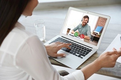 A picture of a woman sitting at a gray wooden table looking at her laptop, where she is video-chatting with a man.