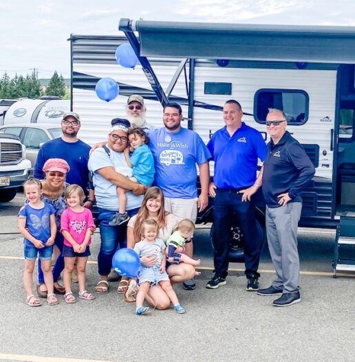 A photo 13 adults and children posing together in a parking lot in front of an RV. Some of the kids have balloons.