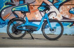 A photo of a blue Elby e-bike in front of a graffitied wall.