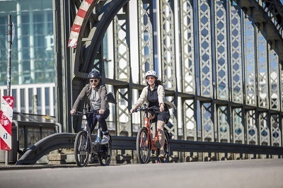 A picture of two people riding electric bikes across a bridge in an urban setting.