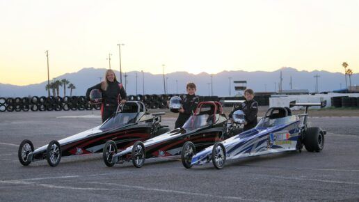 A picture of three people holding helmets and standing next to three drag cars. The sun is setting behind the mountains behind them.