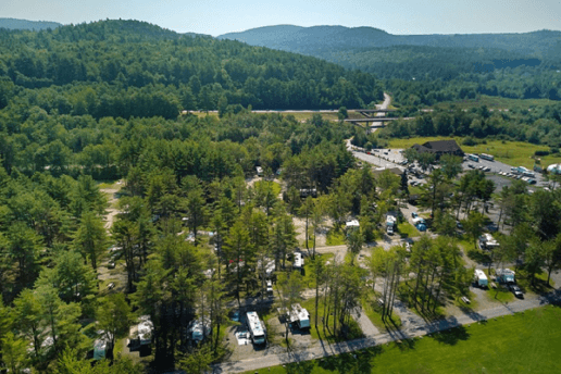 An aerial view of the Lake George Escape RV park. Several RVs are parked beneath tall trees, and rolling hills full of trees are in the background.