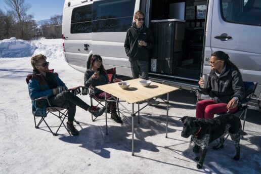 A stock image of four friends camping around a table set outside a Type B motorhome, which is parked on the snow.